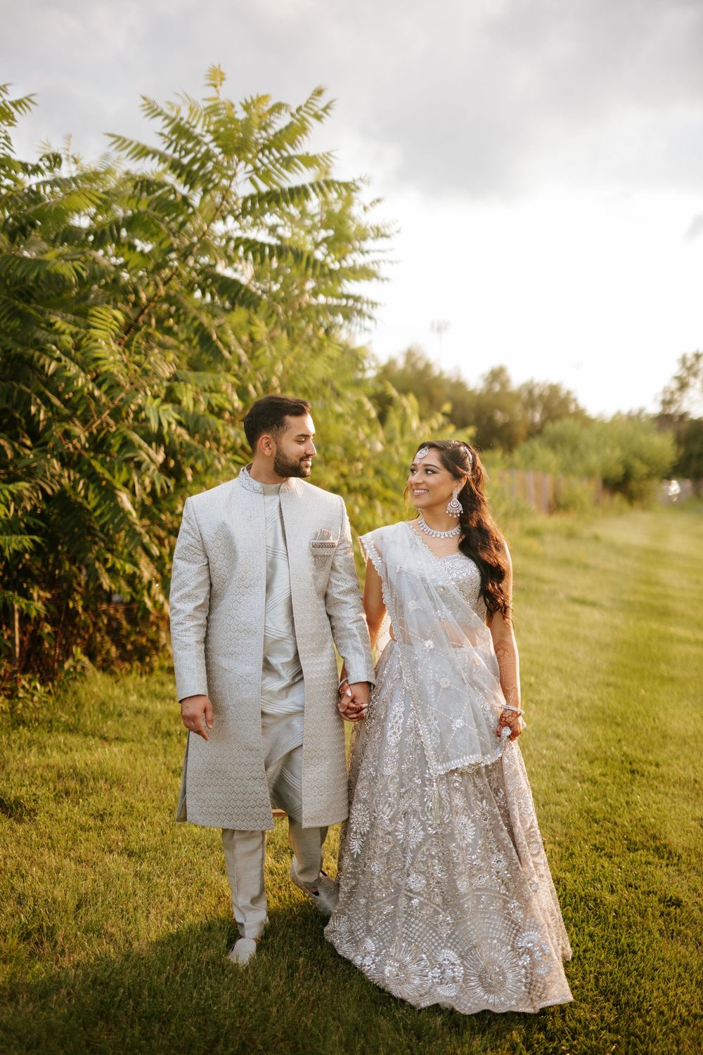 A couple in traditional South Asian wedding attire walk hand in hand through a lush garden setting.