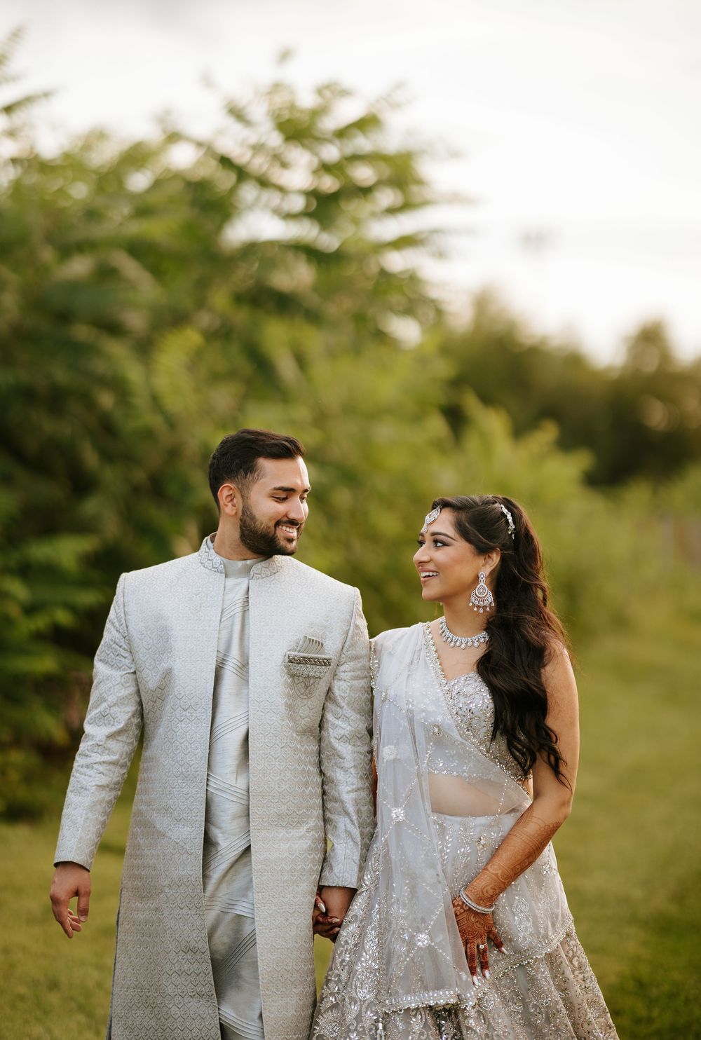 Couple in matching silver outfits walk hand in hand through a lush green garden setting.