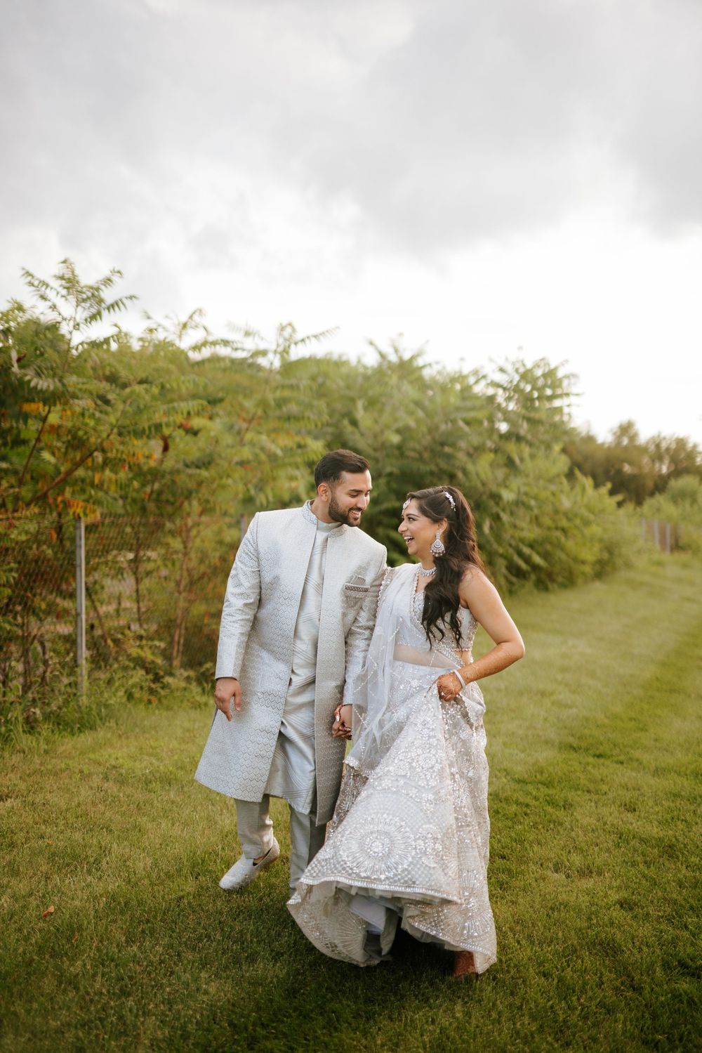 A couple in traditional South Asian wedding attire walk together on a grassy path surrounded by greenery.