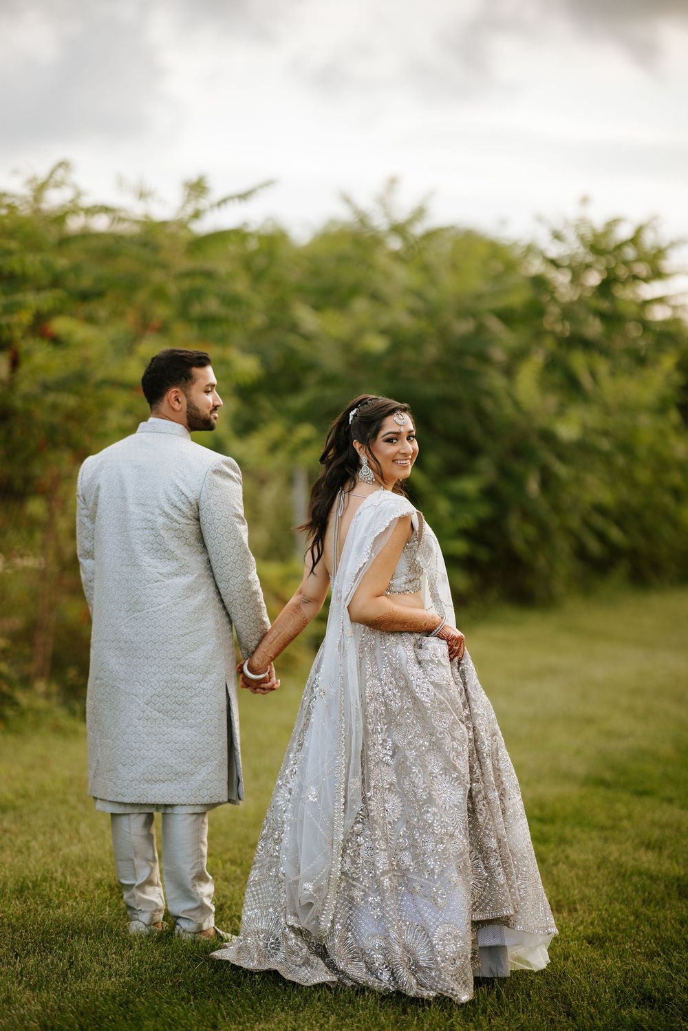 A newlywed couple in silver and white traditional outfits pose in a garden setting.