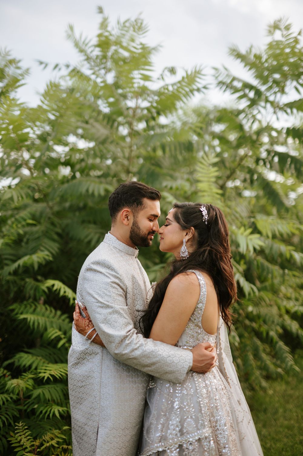A romantic moment between a couple in elegant South Asian wedding outfits against a backdrop of lush foliage.