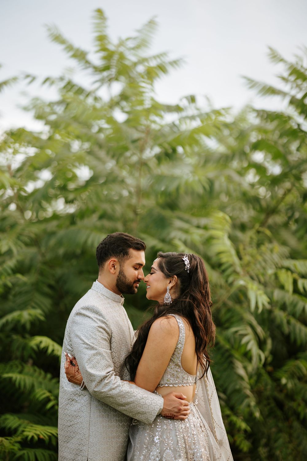 A romantic couple in traditional Indian attire embrace in front of lush green ferns during an outdoor photoshoot.