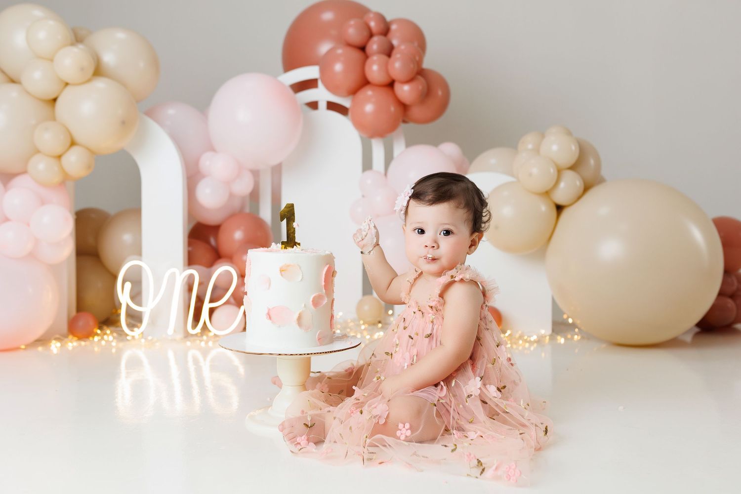 A baby sits among elegant peach and cream colored balloon decorations during a first birthday celebration.