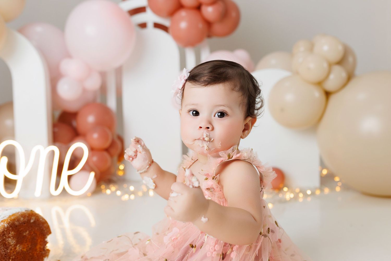 Baby in pink tulle dress sits among pastel balloon decorations and string lights at first birthday celebration.