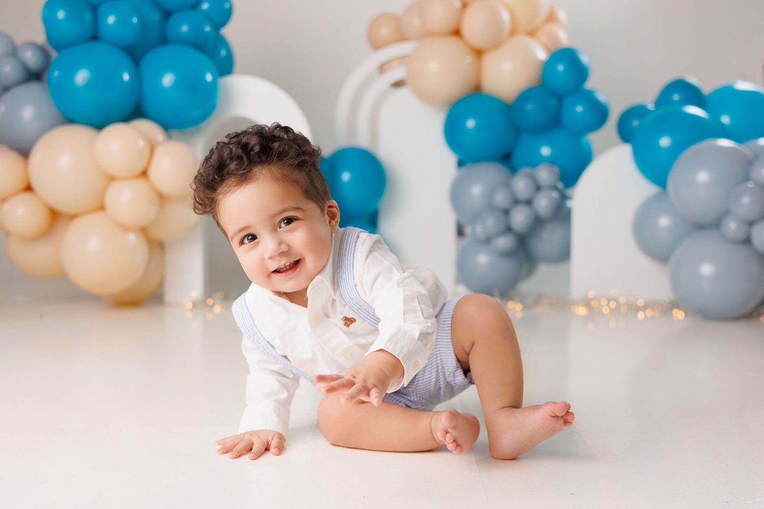 A baby in a white outfit crawls on a white floor with blue and peach colored balloons in the background.