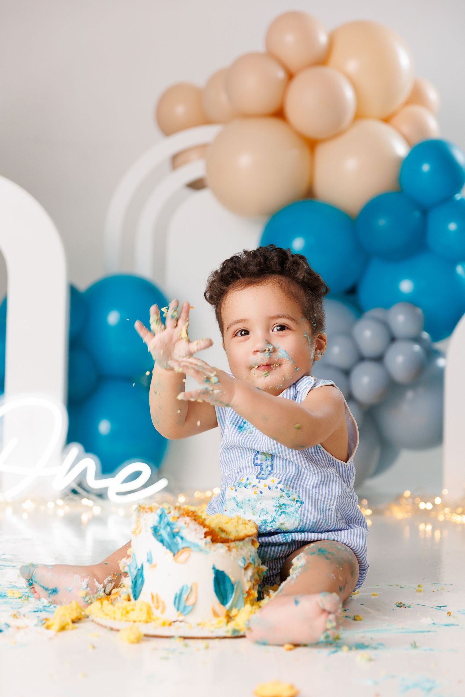 Cake smash photo with blue and peach balloon arch backdrop and twinkling lights for first birthday celebration.