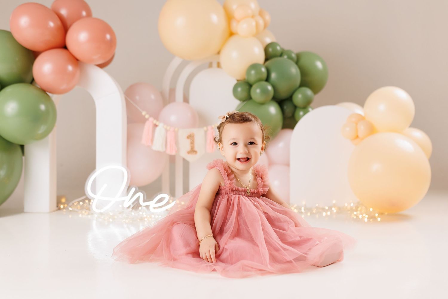 Baby in pink tulle dress sits beneath an elegant balloon arch decorated in soft peach and sage green colors.