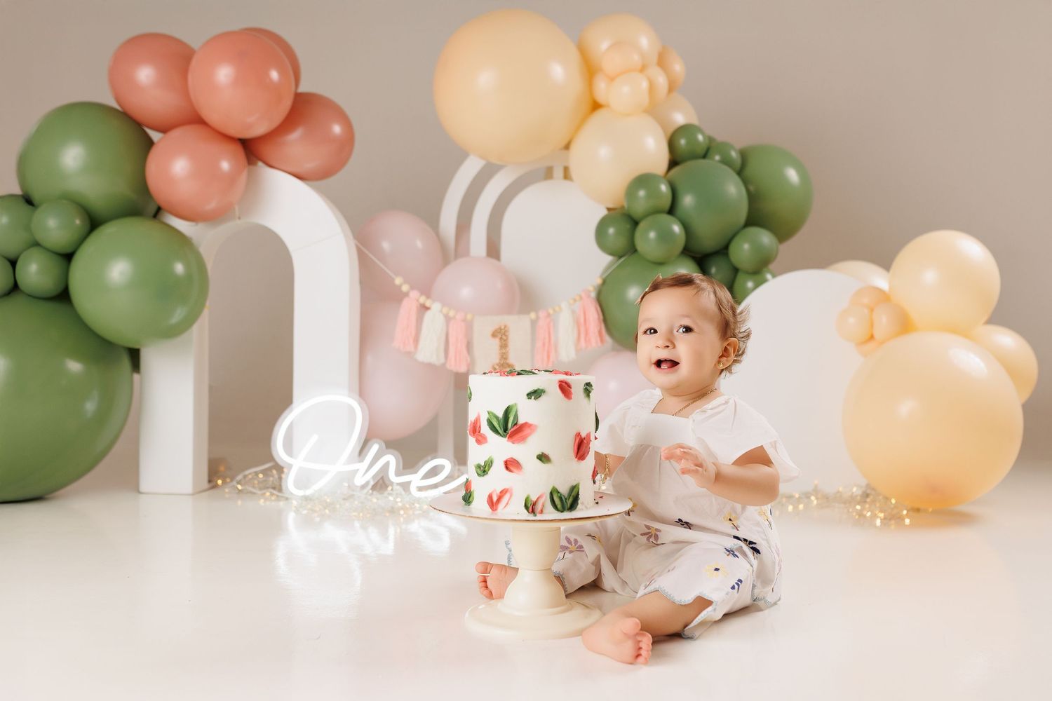 A festive birthday setup with peach, green and cream colored balloons arranged against a white backdrop.