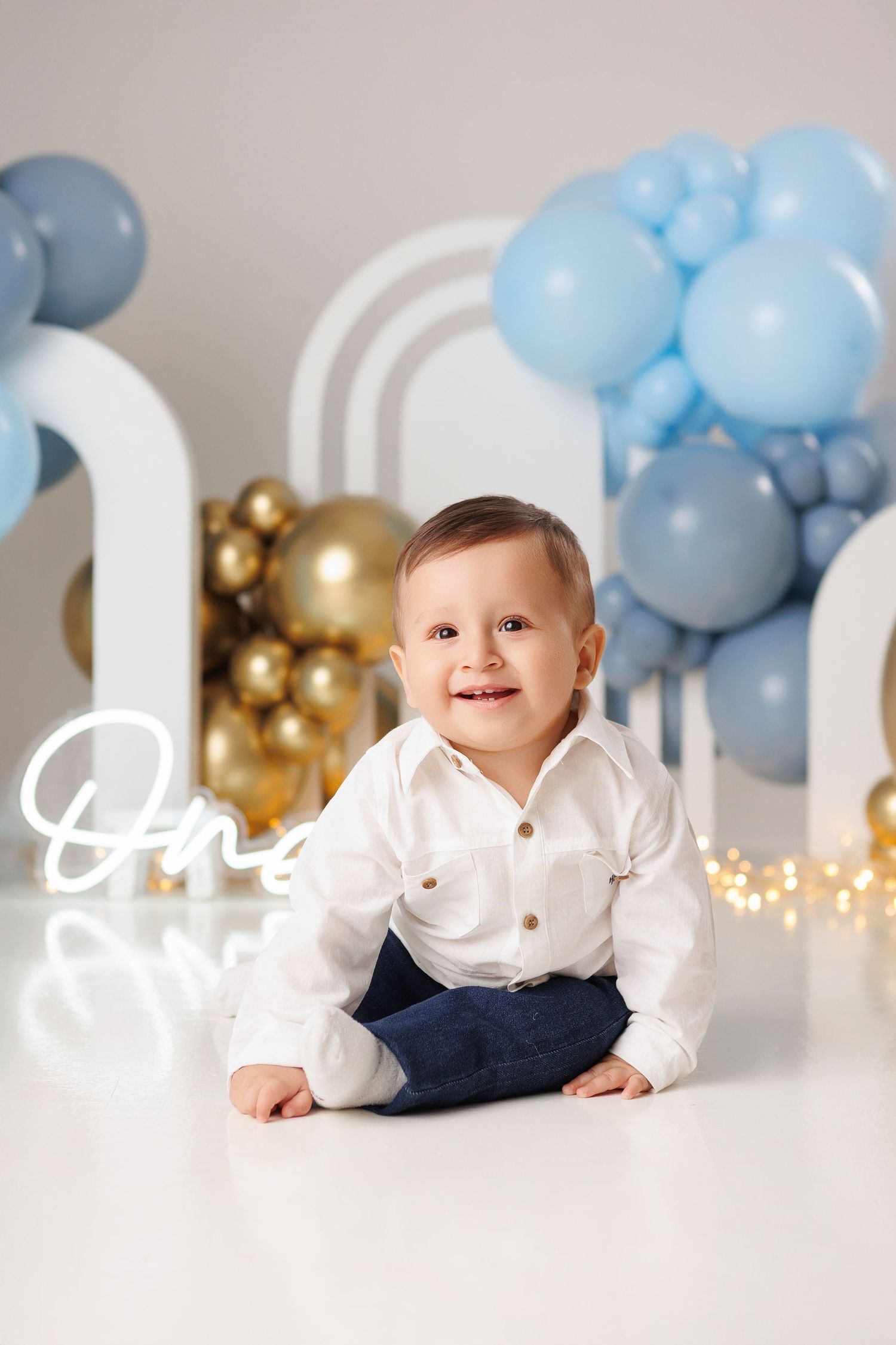 Baby in white shirt and navy overalls lying on floor with blue and gold balloon decorations and white backdrop.