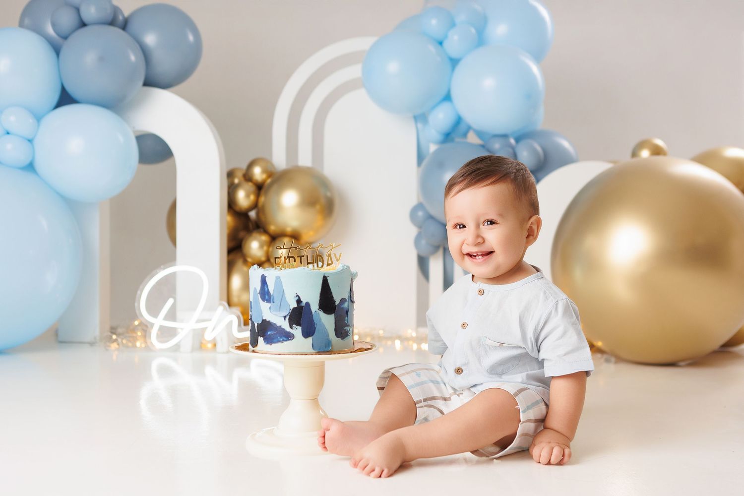 Baby in white shirt sits on floor surrounded by blue and gold balloons during birthday celebration photo session.