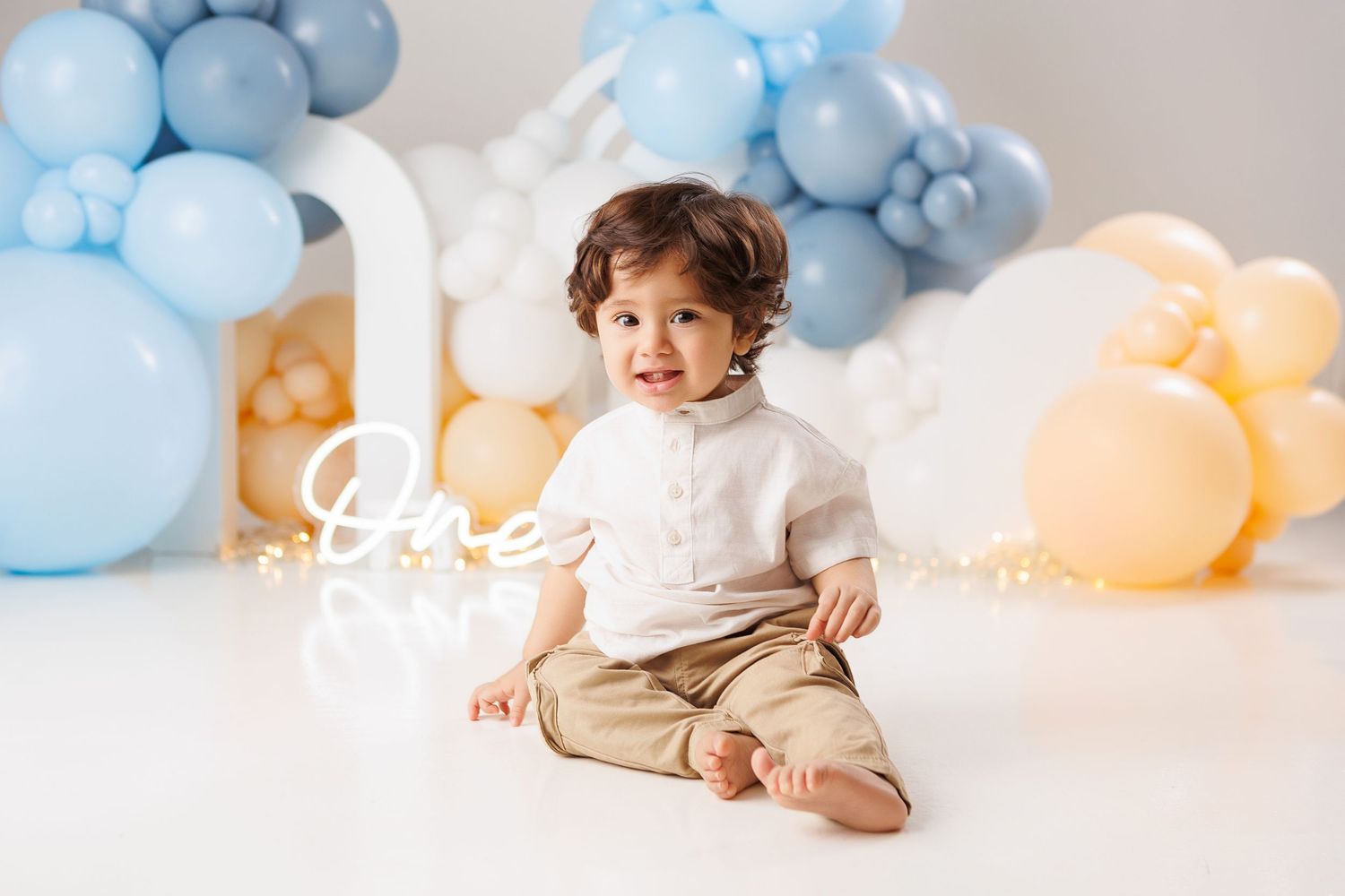 Baby in white outfit sits among blue and peach balloons during first birthday cake smash photo session.