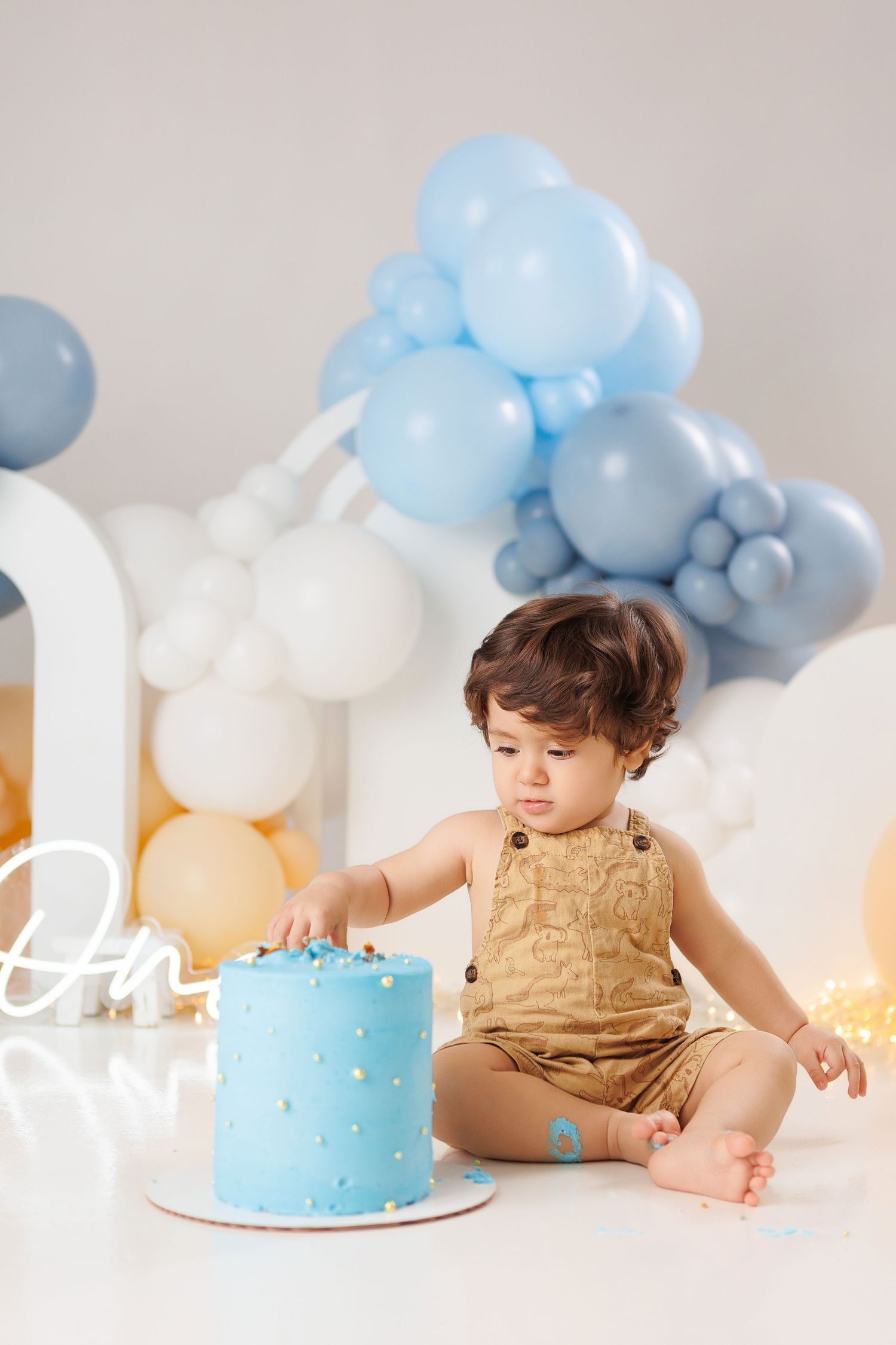 Baby in tan romper sits beside light blue smash cake with white and blue balloon decorations for first birthday celebration.