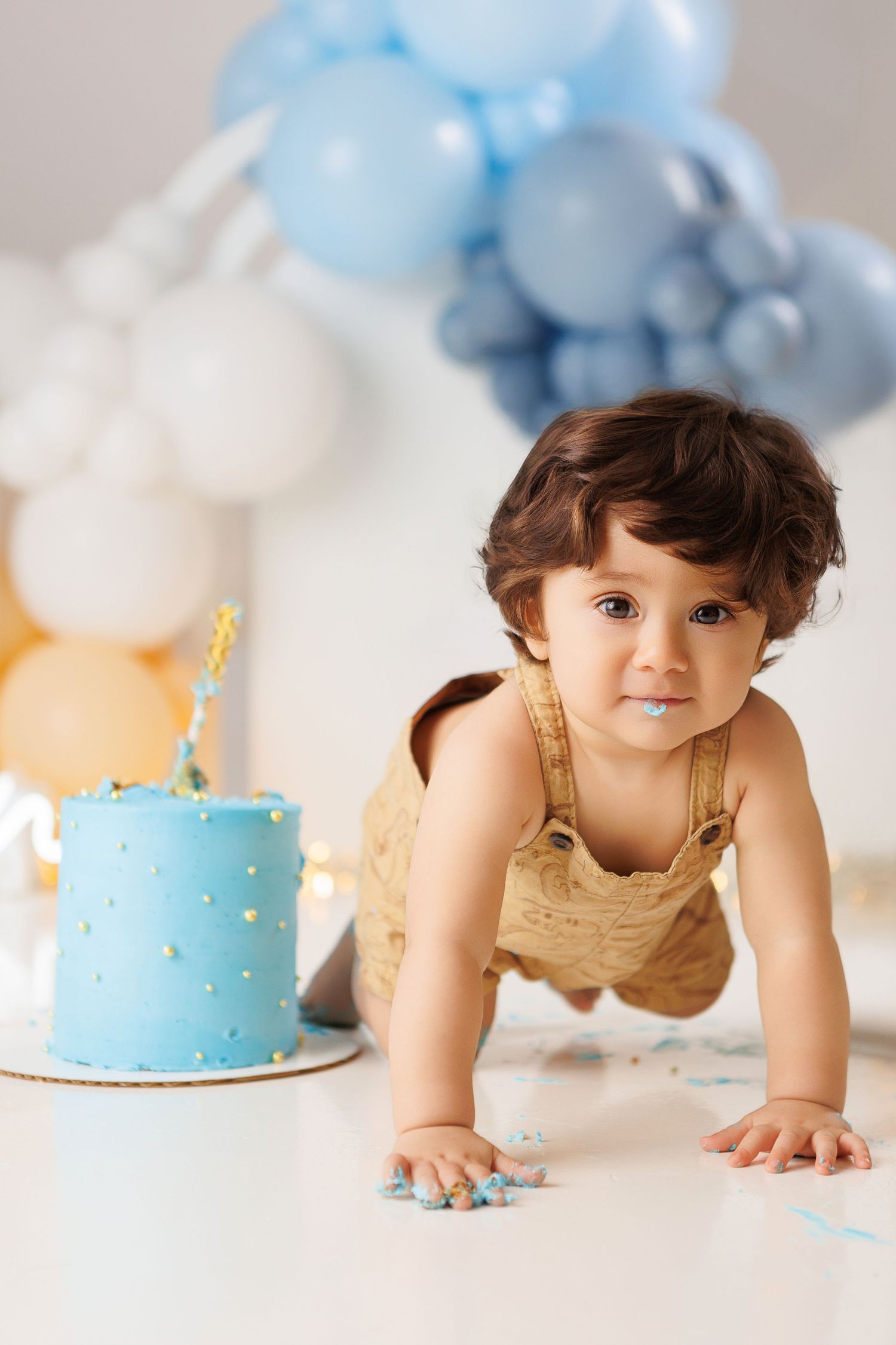 Baby in yellow dress lying on white floor with blue and white balloons and birthday cake during cake smash photo session.