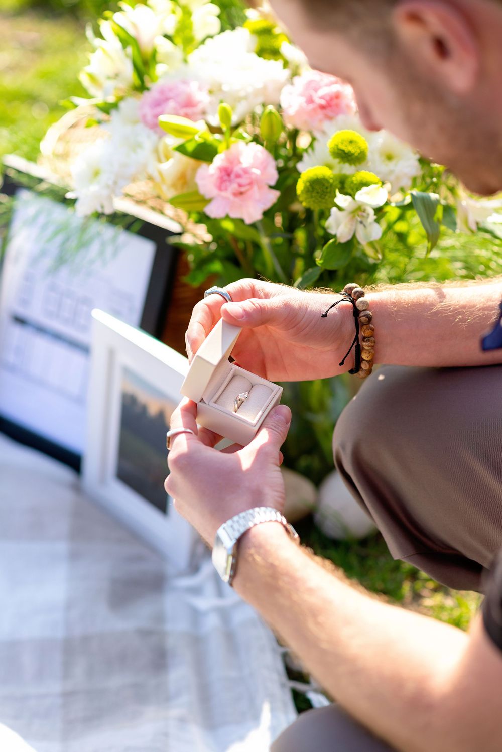 Close-up of hands opening a small ring box with pink and white flowers in the background during a proposal in Bayfield.