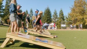 Outdoor event on sunny grass field with people participating in a cornhole tournament.