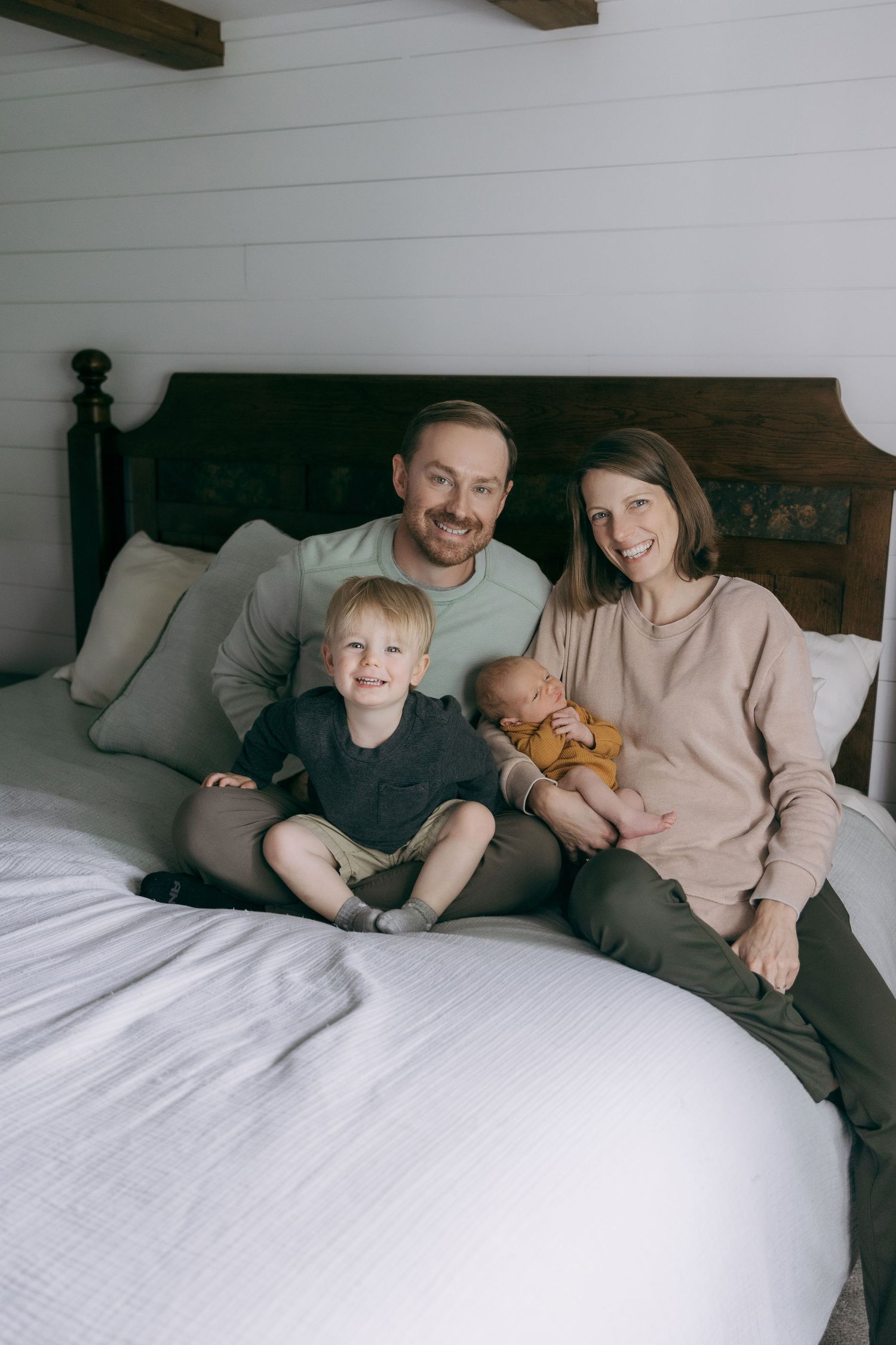 A family relaxes together on a cozy bed with white linens and wooden beam details visible above.