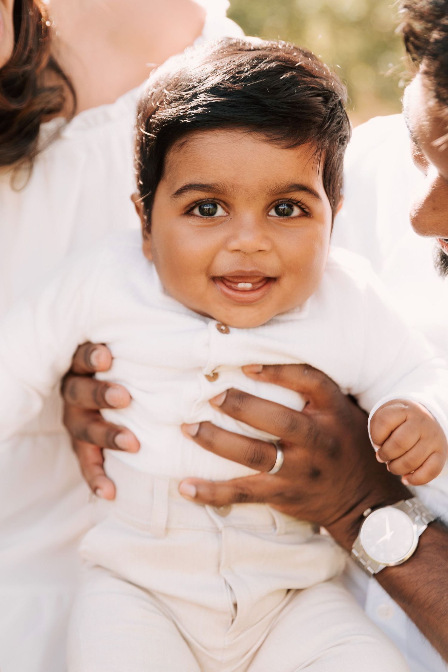 A heartwarming outdoor family photoshoot in a sunlit wooded area with everyone wearing white clothing.