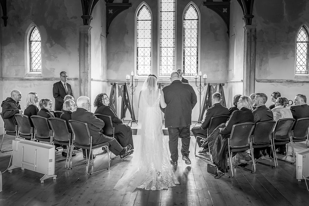 Black and white panorama of a wedding ceremony in a historic church with arched windows and wooden pews.