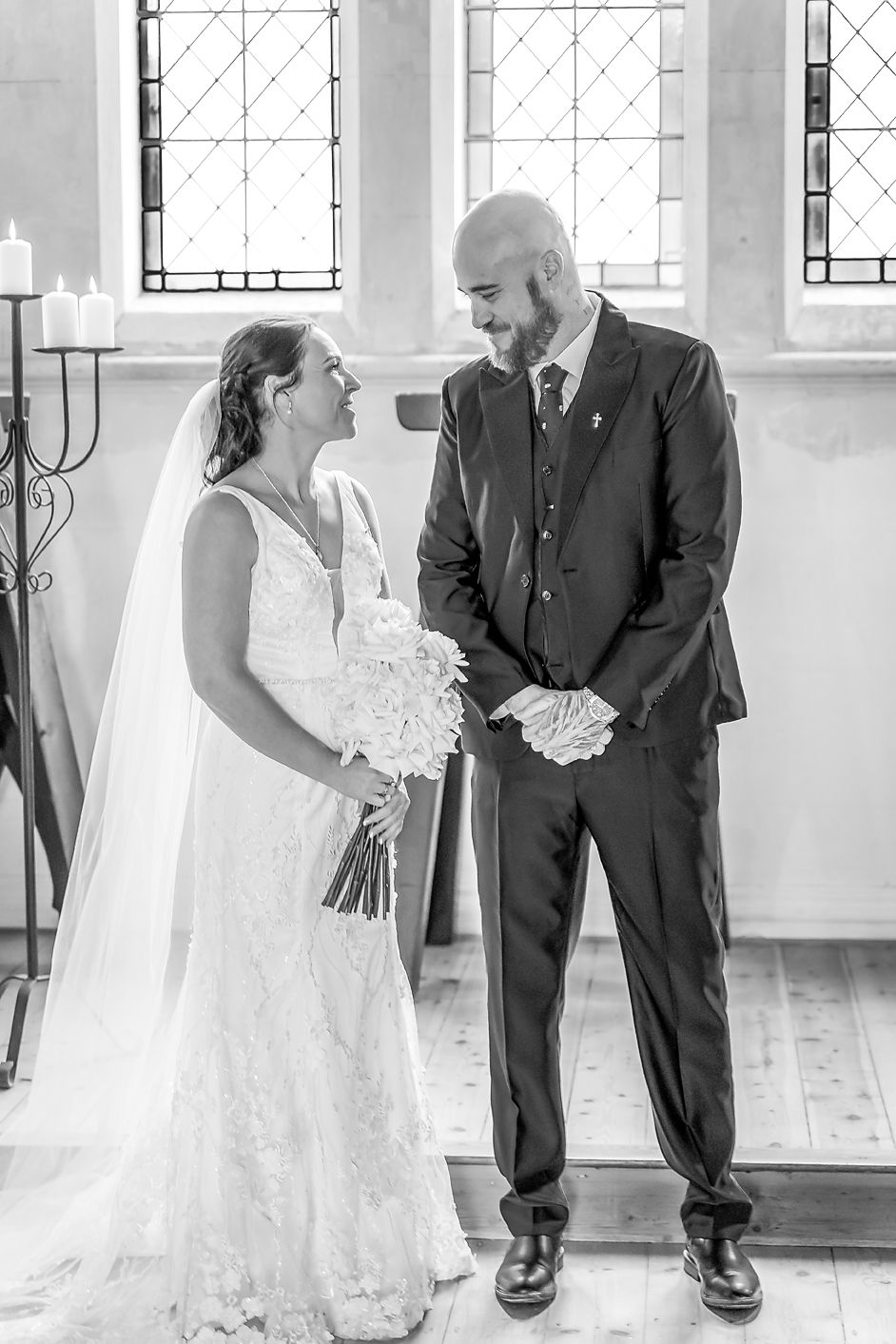 Wedding couple stands in front of sunlit church windows in elegant black and white portrait.