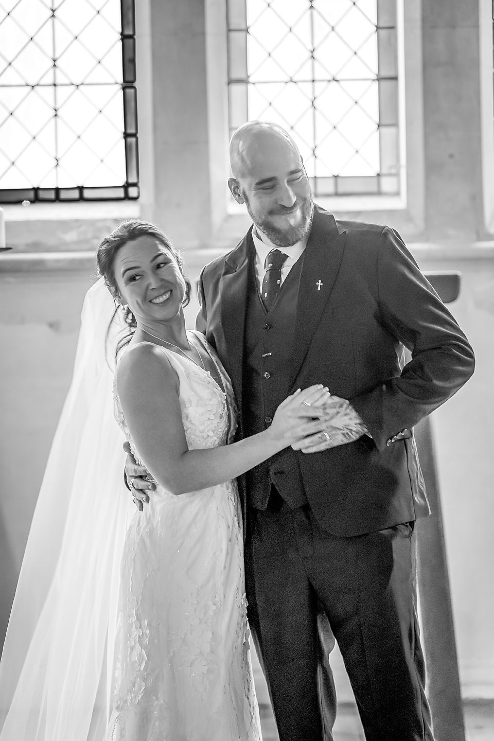 A joyful black and white wedding portrait of a couple sharing a candid moment by a church window.