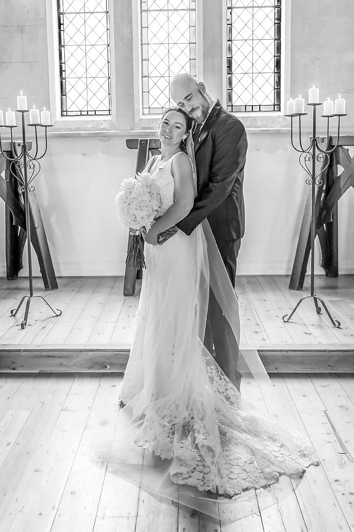 A romantic black and white wedding photo of a couple embracing in a church with candelabras and stained glass windows.