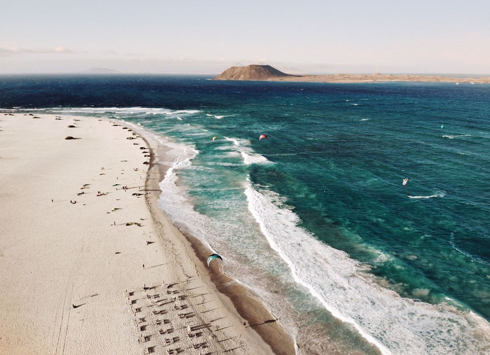Aerial drone view of the turquoise ocean waves hitting the white sands of Grandes Playas with Los Lobos island in the distance