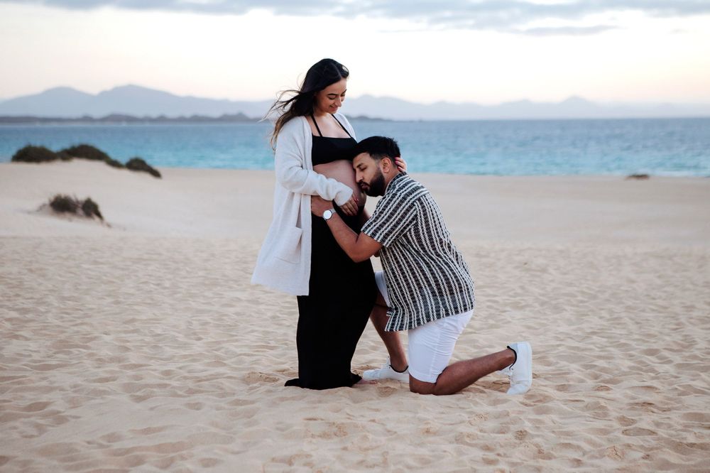 Man kneeling to kiss his partner's pregnant belly in the Corralejo dunes at sunset - Emotional maternity photography in Fuerteventura
