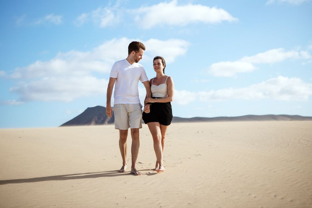Couple walking hand-in-hand through the vast white sand dunes of Corralejo with mountains in the background - Fuerteventura couple session