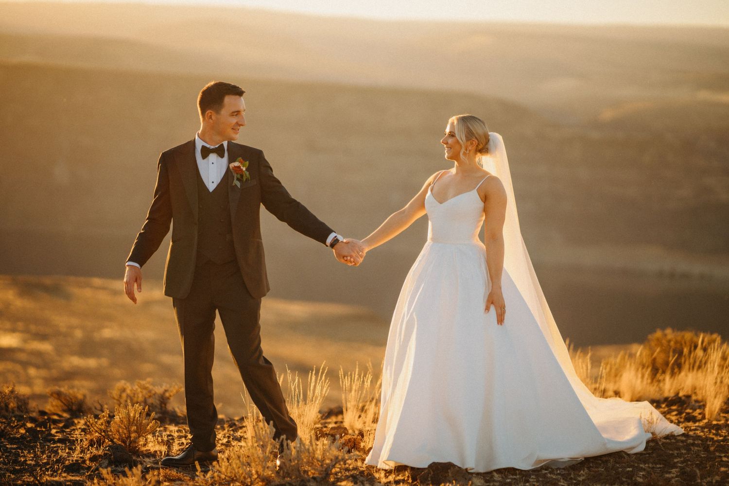 A couple in wedding attire stands silhouetted against a golden sunset on a hilltop overlooking a vast landscape.