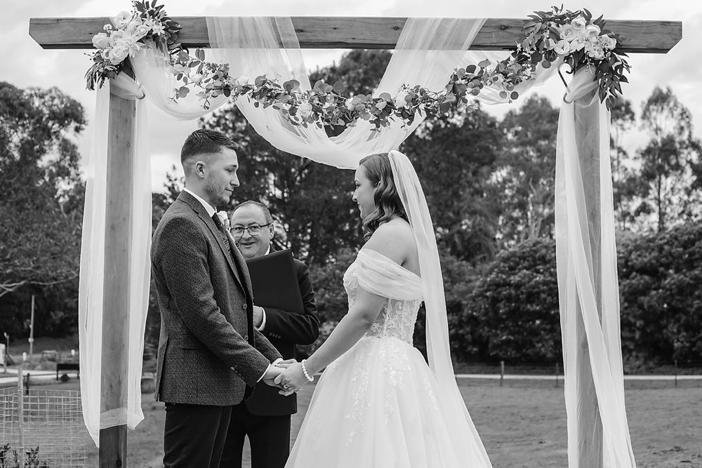 A black and white wedding ceremony under a decorated pergola with white fabric and floral arrangements.