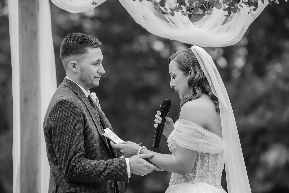 Black and white wedding ceremony photo showing couple exchanging vows under elegant outdoor floral arch.