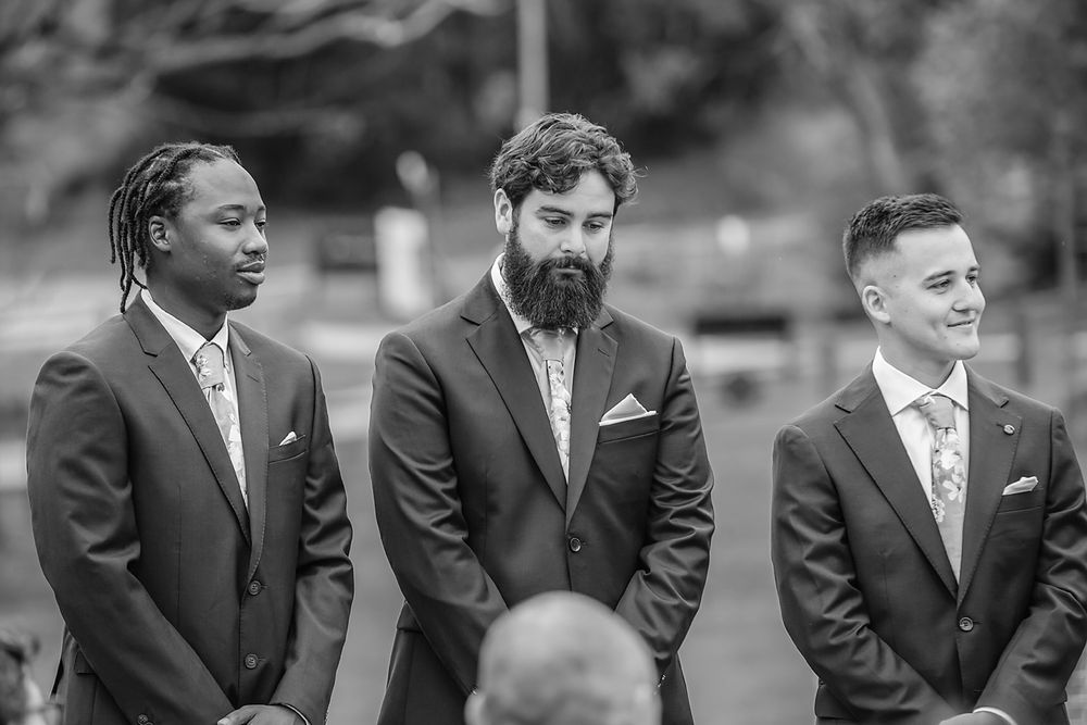 A group of groomsmen in black suits and ties stand together smiling during an outdoor wedding ceremony.