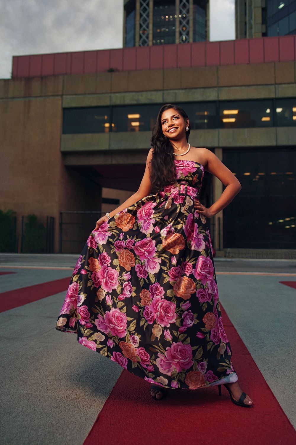 A woman in a stunning floral evening gown with pink and black roses poses on a red carpet outside a building.