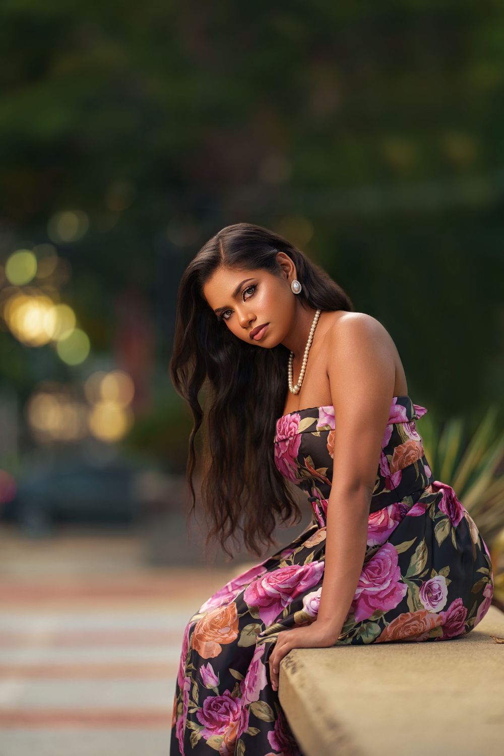 A floral maxi dress with pink roses is showcased in an outdoor portrait with bokeh lighting effects in the background.