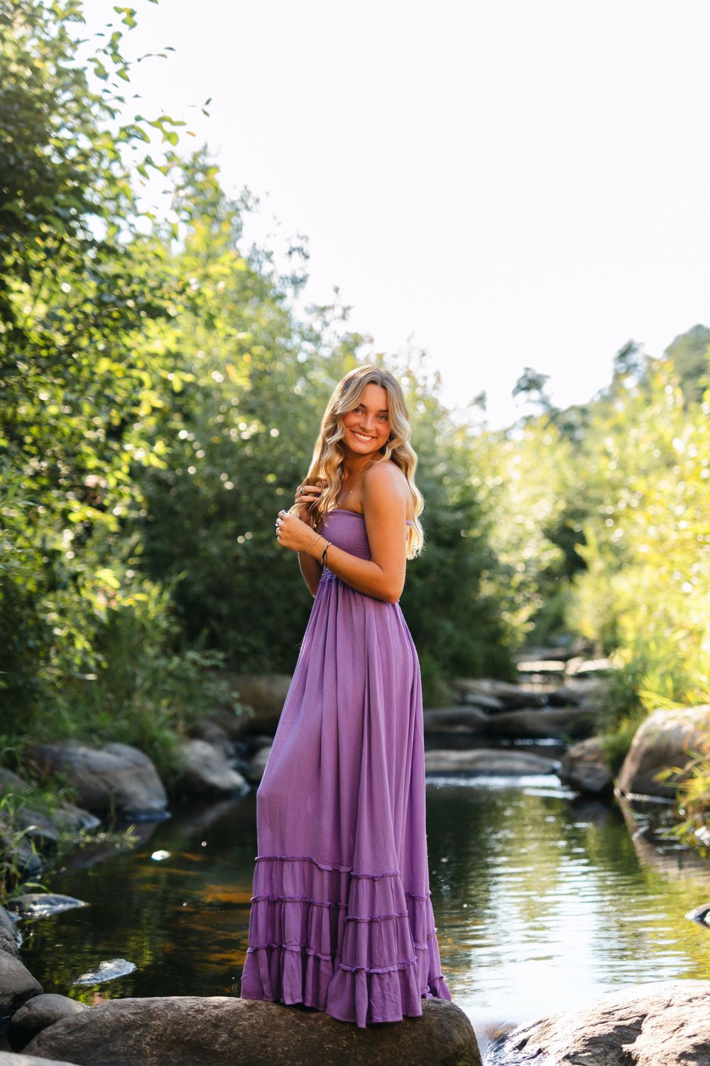 A purple maxi dress flows gracefully as someone poses on rocks near a tranquil stream in dappled sunlight.
