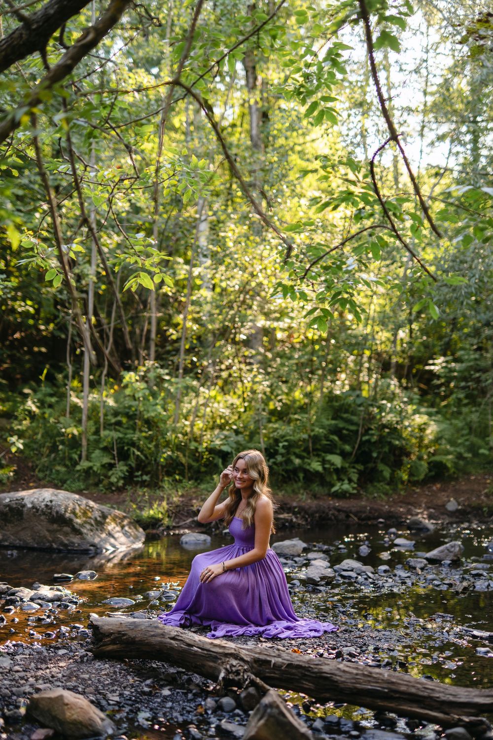 A flowing purple dress catches sunlight in a forest creek setting with rocks and fallen logs.