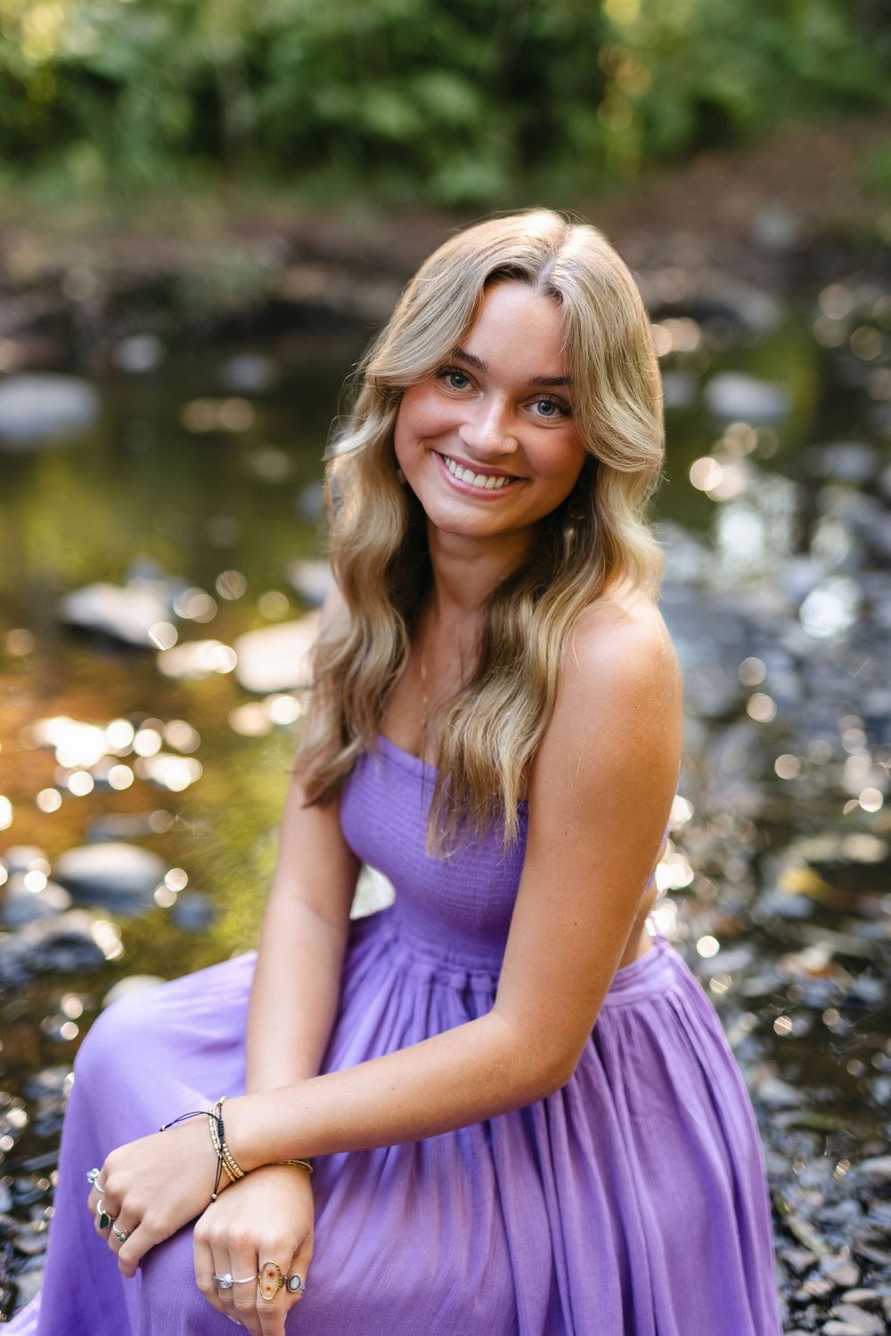 A series of outdoor portraits showing someone in a flowing purple dress sitting by a stream with rocks and natural lighting.