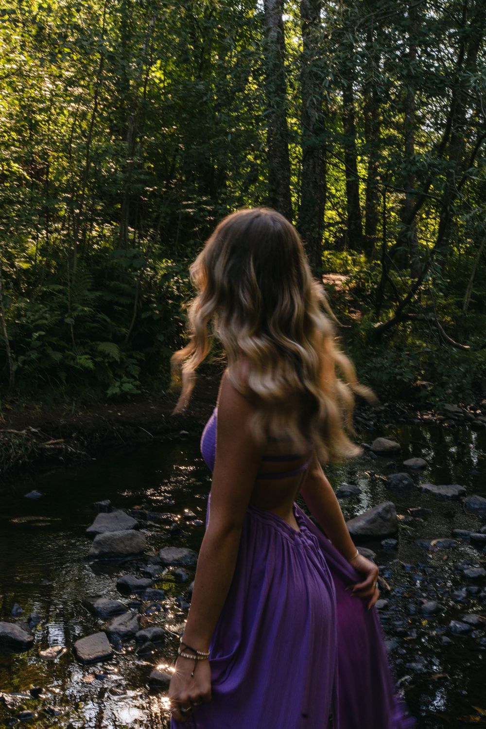 A woman in a flowing purple dress stands by a stream in a sunlit forest setting.