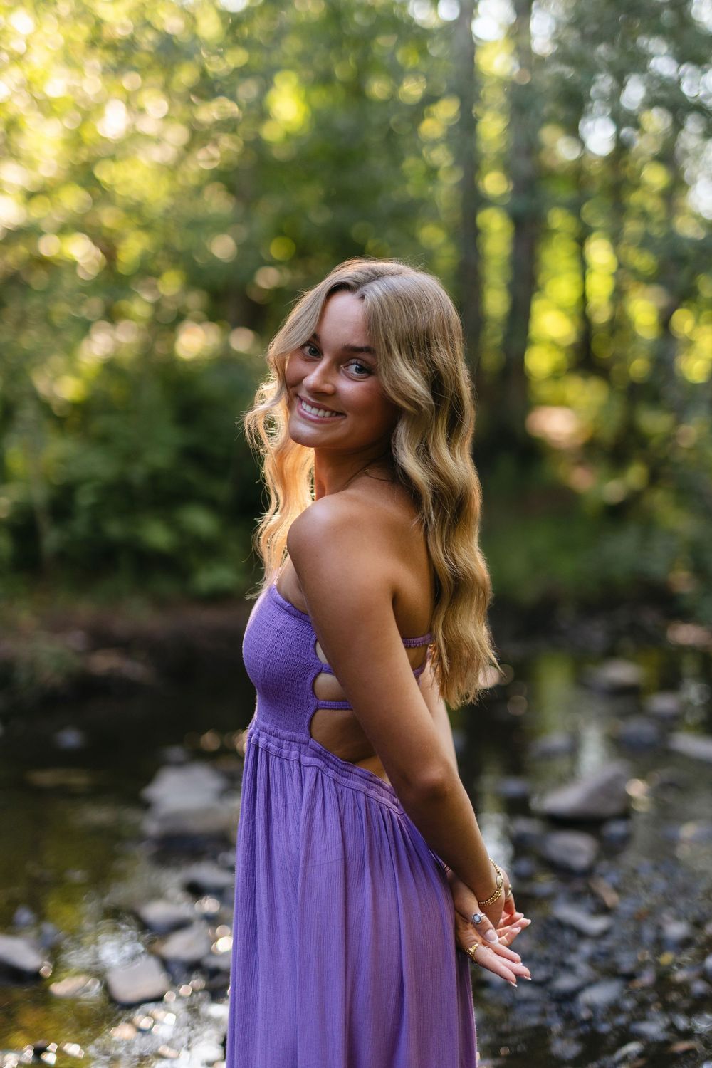 A purple cutout dress sparkles in golden sunlight by a forest stream.