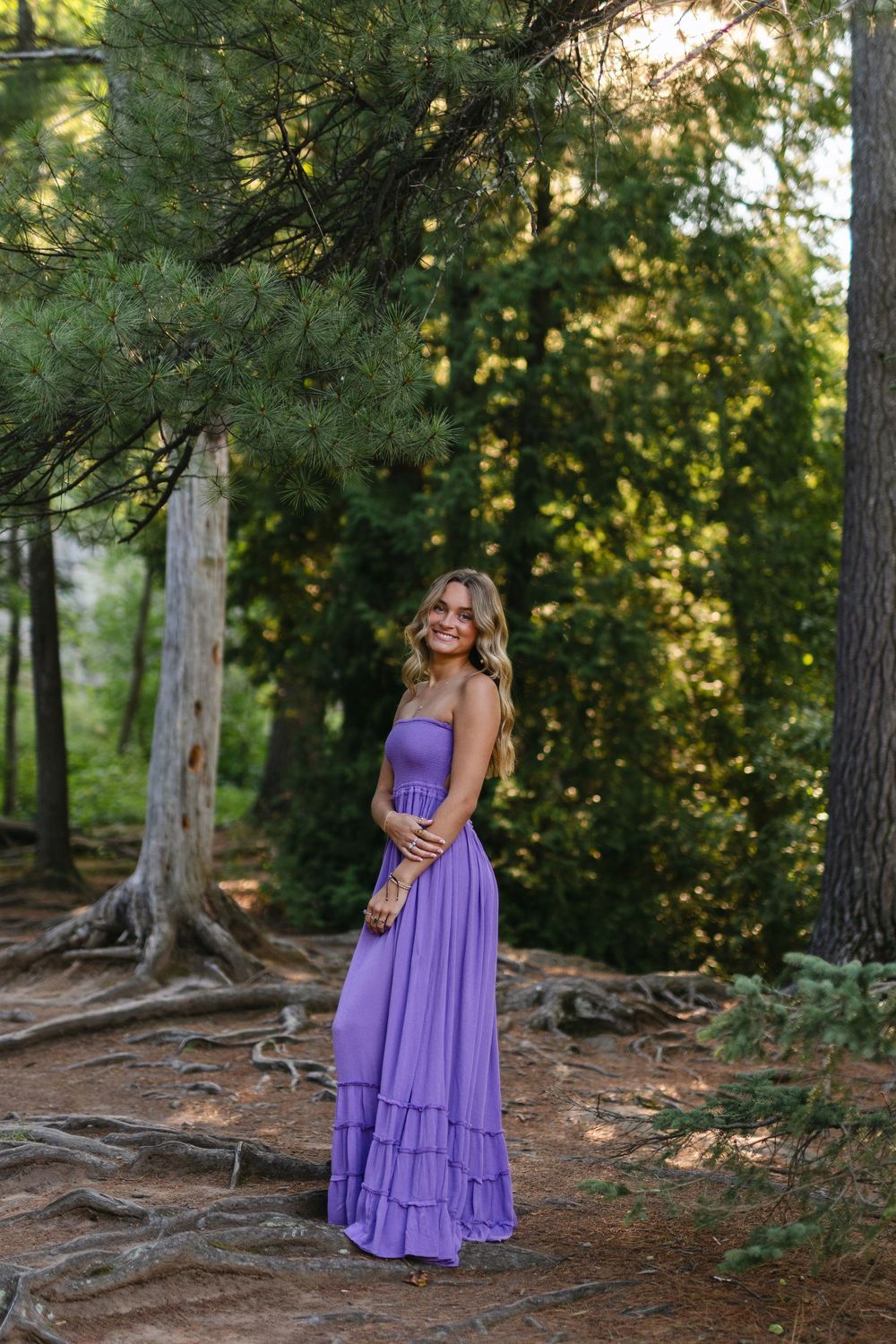 A purple maxi dress flows elegantly against a backdrop of tall pine trees in a forest setting.