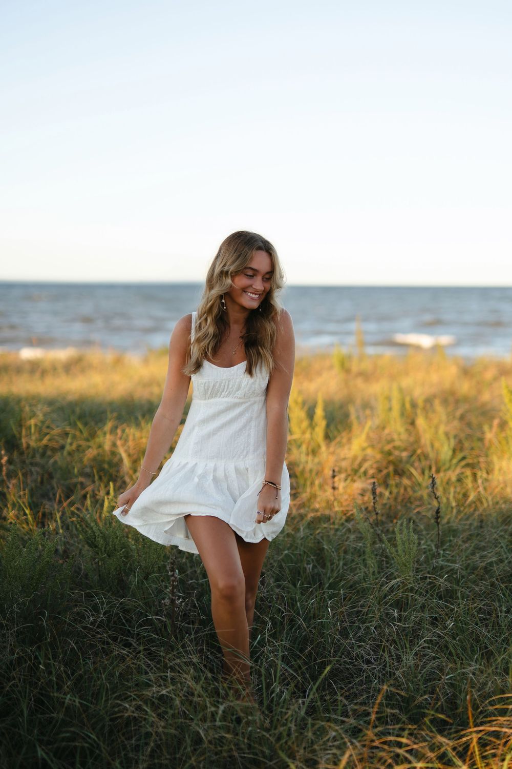 A person in a white summer dress poses playfully in a field of tall grass at sunset near the ocean.