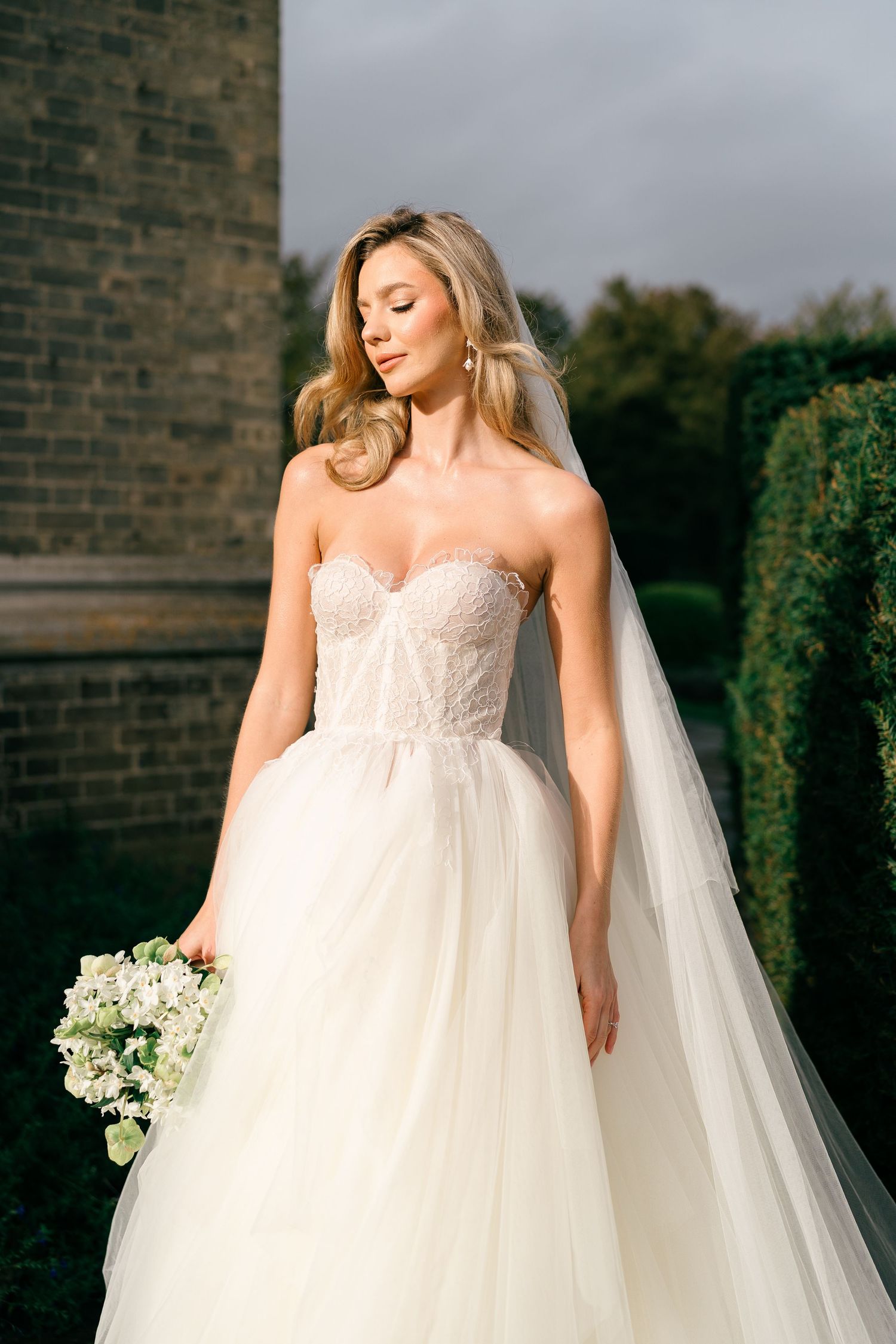A bride in a flowing empire waist wedding gown with lace bodice and long veil poses in front of a brick wall.