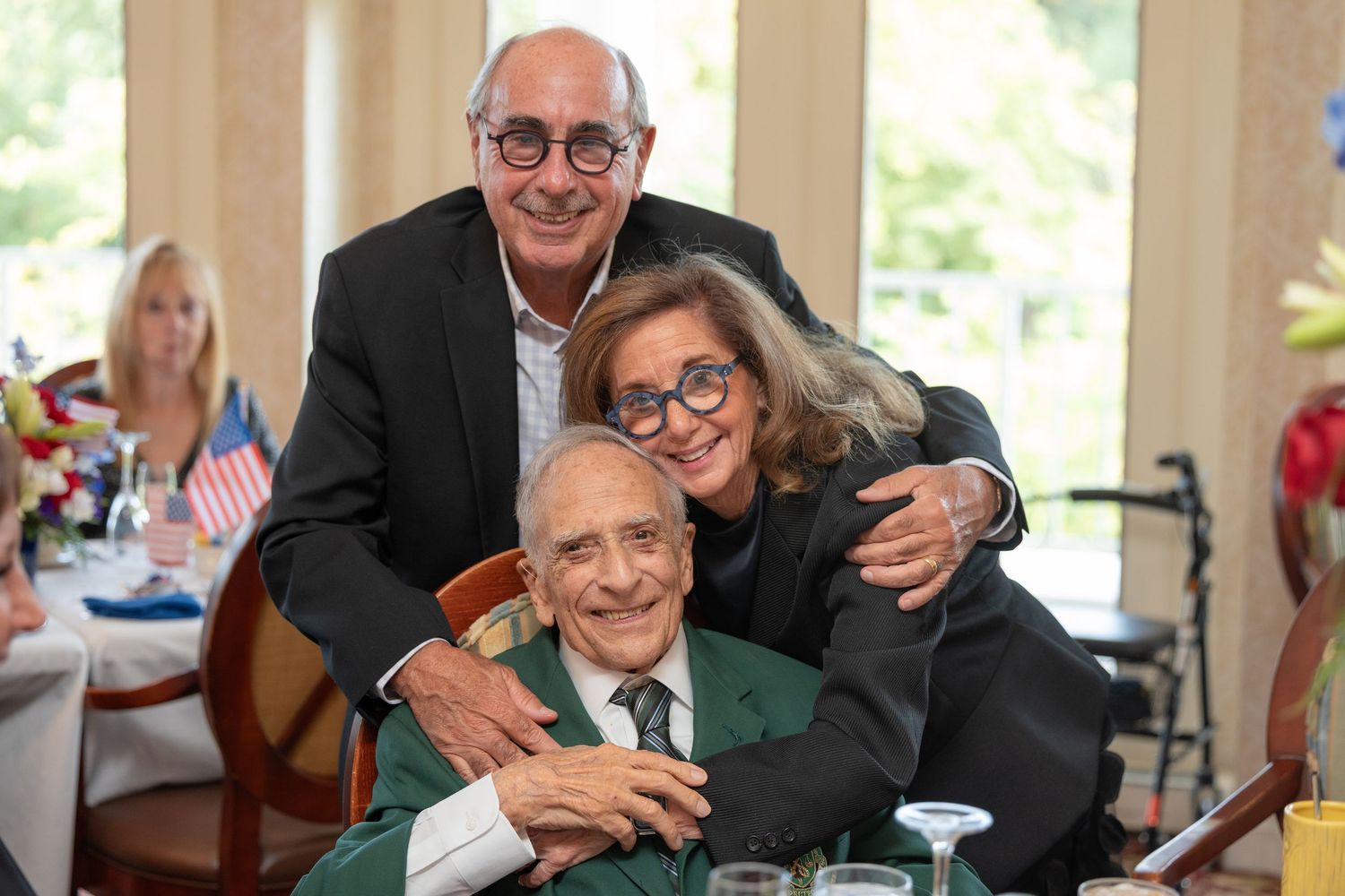 Three people share a warm moment at a celebration event with American flags visible in the background.