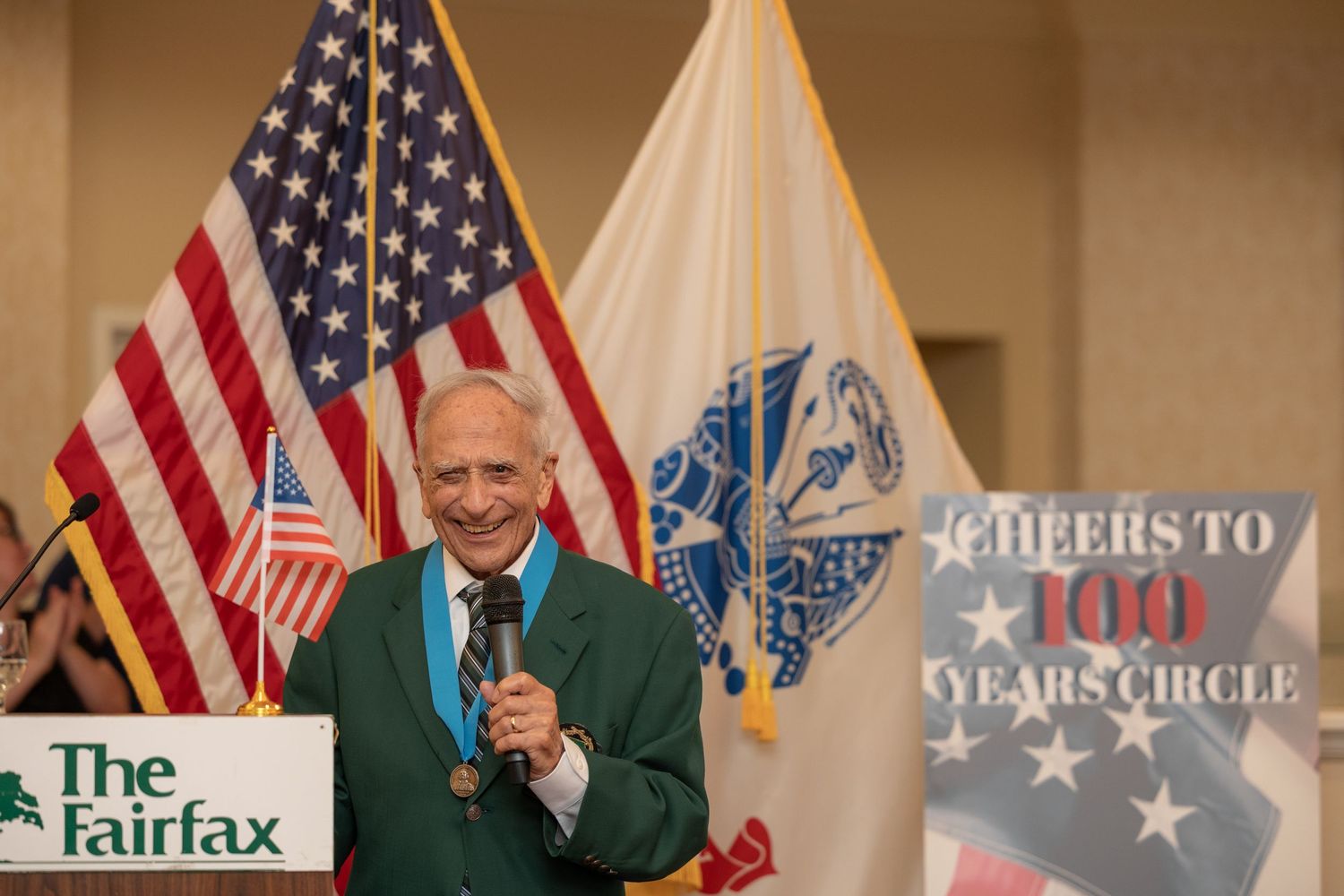 Person in green blazer speaks at podium with American flag and event banner visible behind them.