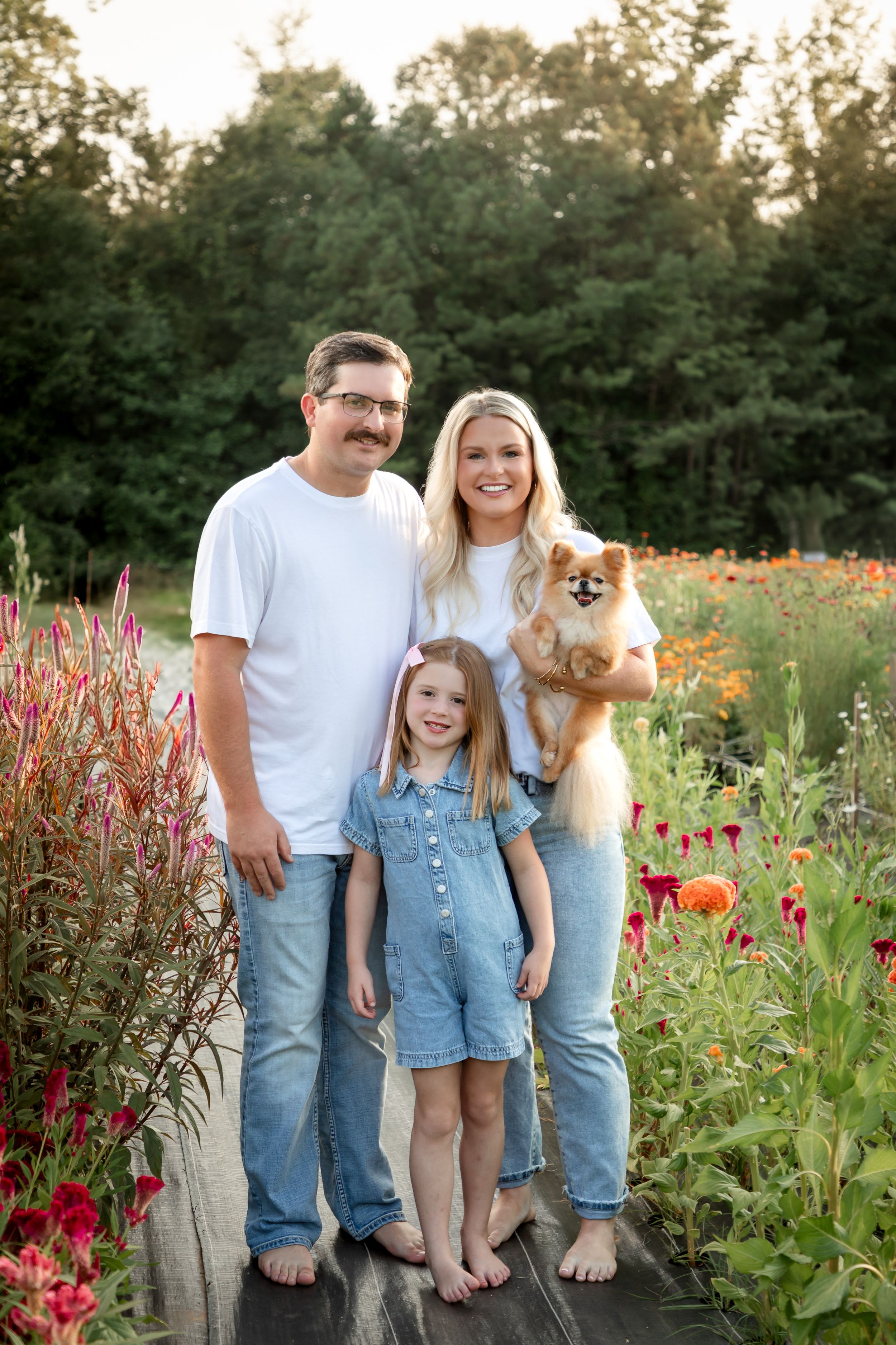A family poses barefoot on a wooden deck surrounded by vibrant wildflowers and greenery while holding a Pomeranian dog.