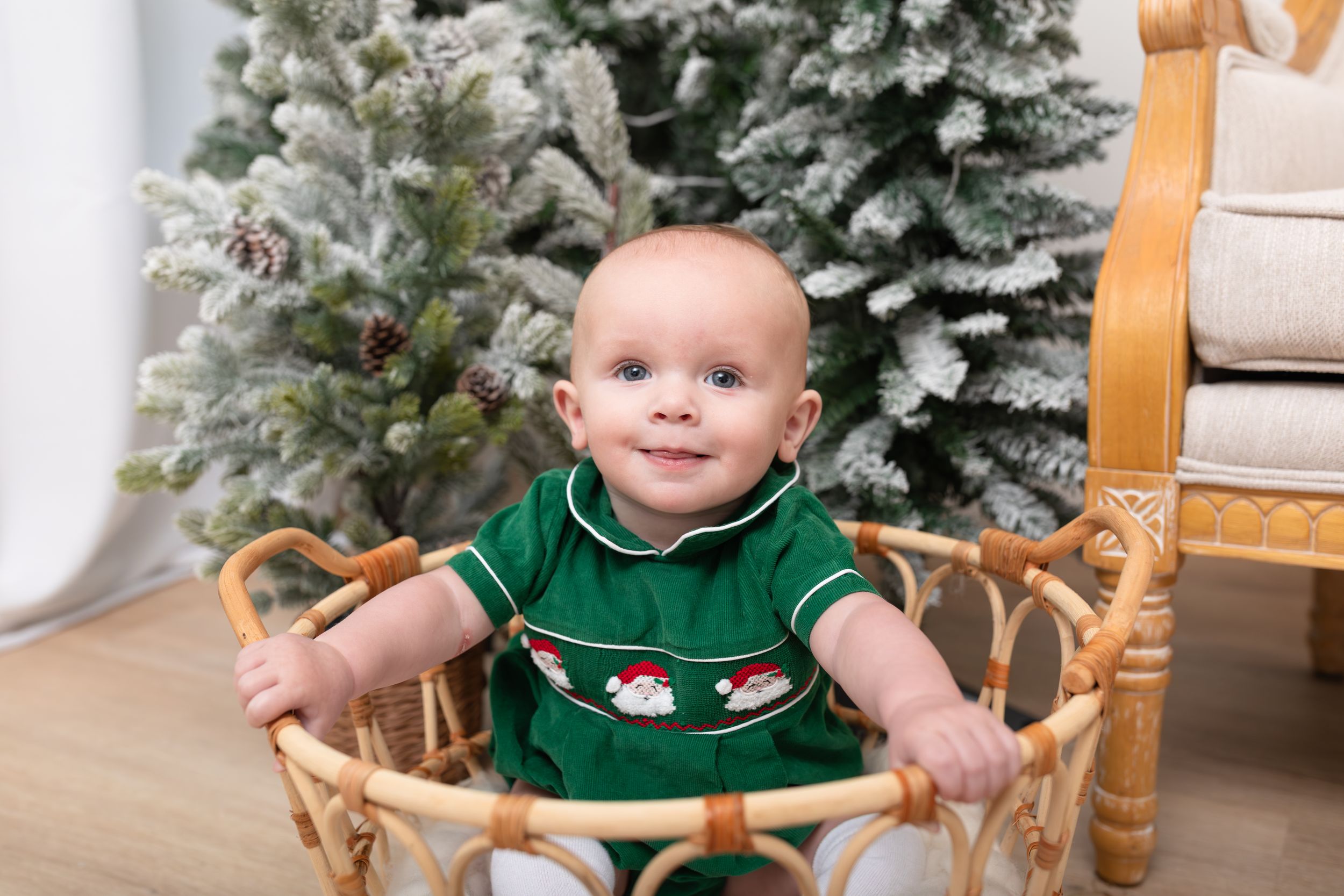 A baby in a green Christmas outfit sits in a wooden chair near a decorated holiday tree.