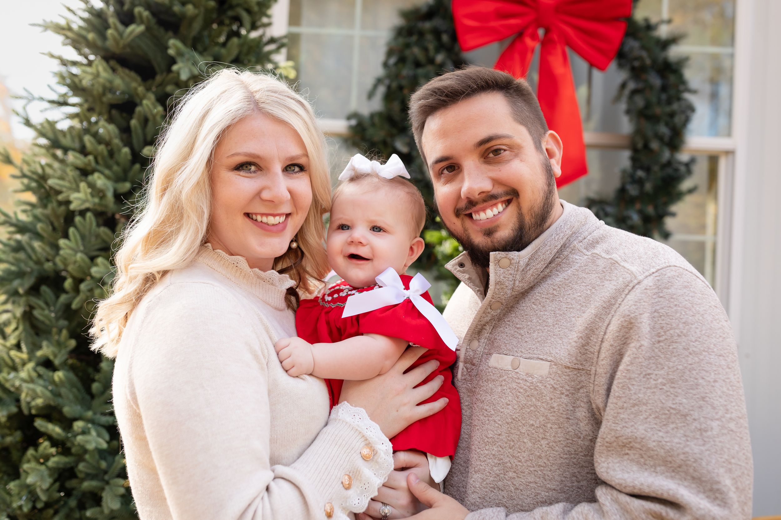 Family poses together outdoors by evergreen tree and red holiday bow during Christmas season.