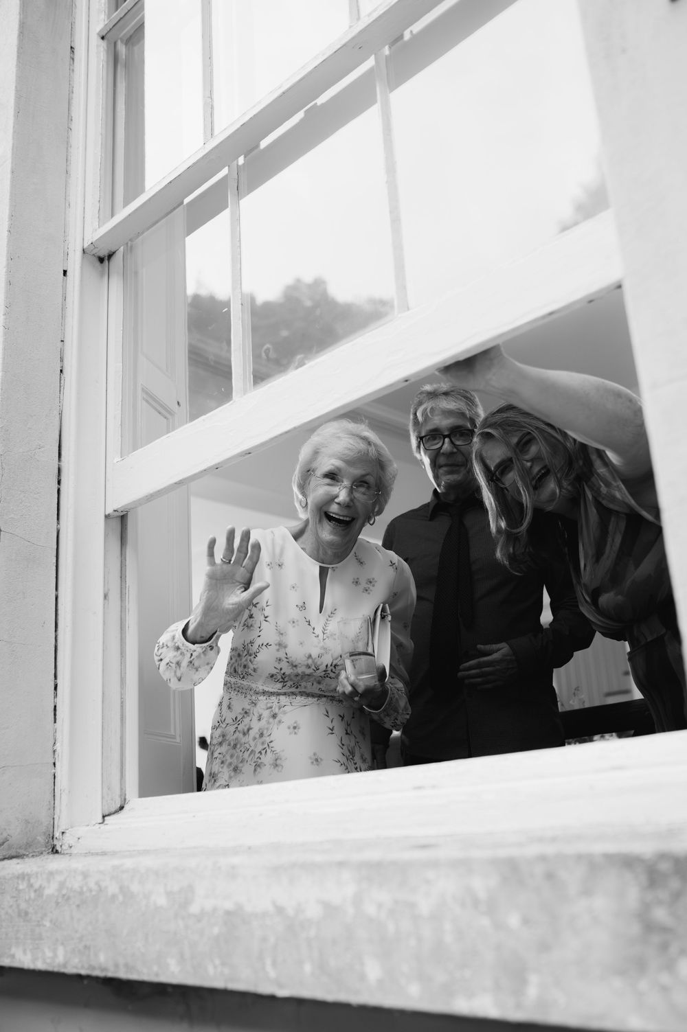 A black and white photograph shows people peeking and waving through windows of what appears to be an old building.