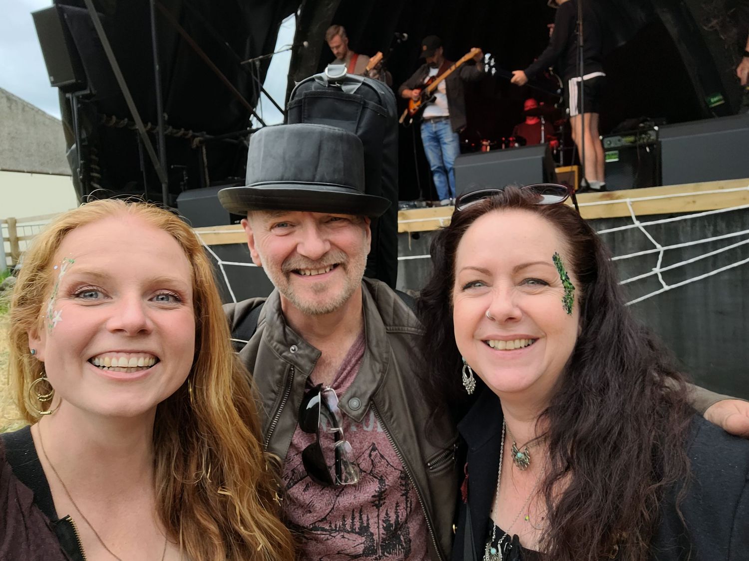 Three people pose together at an outdoor concert venue with a stage and musicians visible in the background.