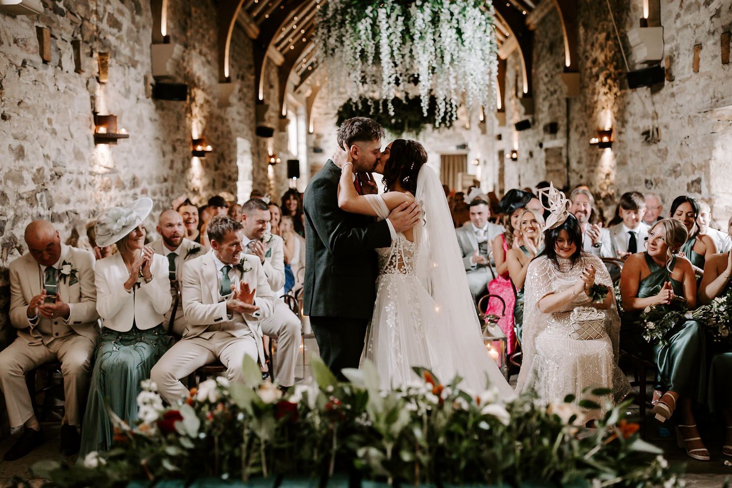 A romantic wedding ceremony in a rustic stone venue with hanging greenery and guests seated on both sides.